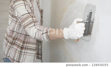 Unknown female construction worker wearing protective gloves applying plaster, smoothing wall surface during home renovation work 129438355