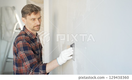Construction worker man wearing gloves and holding a putty knife is applying plaster on a white wall, performing home renovation and improvement Construction worker man wearing gloves and holding a putty knife is applying plaster on a white wall, performing home renovation and improvement 129438360