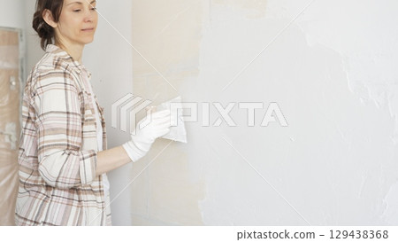 Woman applying plaster to wall with putty knife, wearing protective gloves and creating smooth surface during home renovation work Woman applying plaster to wall with putty knife, wearing protective gloves and creating smooth surface during home renovation work 129438368