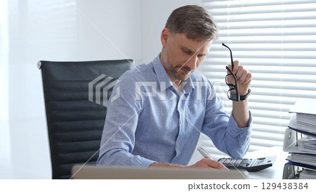 Adult businessman wearing blue sleek shirt and glasses, is thinking and concentrating while working at the desk in a bright, modern office, demonstrating professionalism and efficiency. Business 129438384