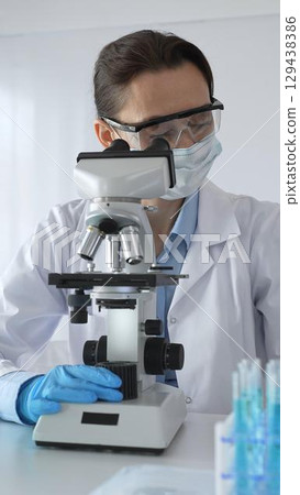 Female scientist examining microscope slide, wearing protective gear in sterile laboratory, researching pharmaceutical breakthrough with blue tinted test tube specimens 129438386