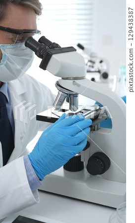 Man scientist wearing protective googles and blue gloves, is using microscope analyzing samples in modern laboratory doing scientific research. Medicine and science concept 129438387