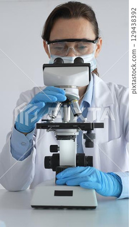 Female scientist examining microscope slide, wearing protective gear in laboratory, researching pharmaceutical breakthrough with blue tinted test tube specimens 129438392