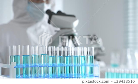 Medical scientist wearing protective suit using microscope while analyzing samples, conducting research in laboratory environment. Close up of test tubes with blue liquid 129438510