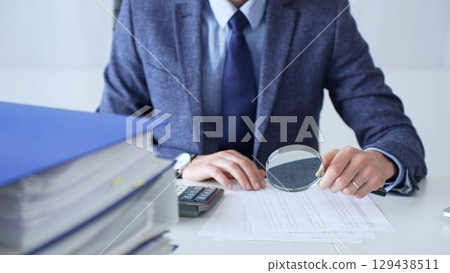 Professional businessman examining financial documents, magnifying glass in hand, calculator positioned on sleek desk workspace. Audit and taxes in business 129438511