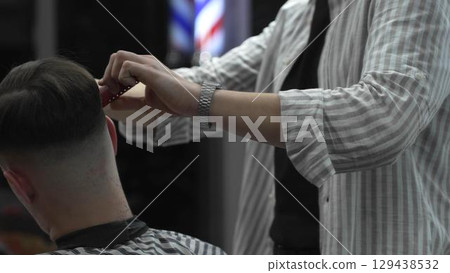 Skilled barber trimming customer's hair using professional scissors and comb, working in contemporary barbershop with vintage pole visible near workspace 129438532