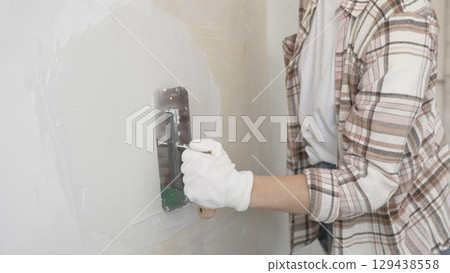 Unknown female construction worker wearing protective gloves applying plaster, smoothing wall surface during home renovation work 129438558