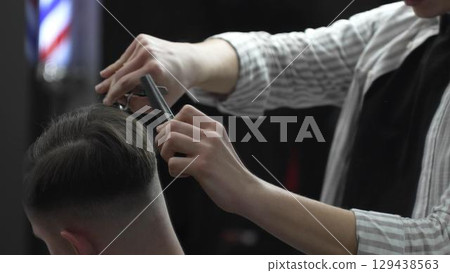 Skilled barber trimming customer's hair using professional scissors and comb, working in contemporary barbershop with vintage pole visible near workspace 129438563