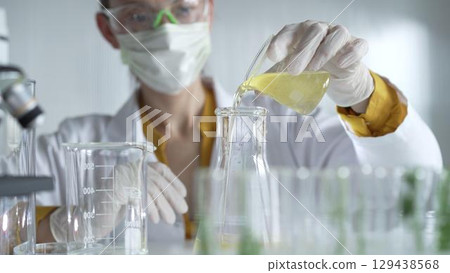 Female scientist researcher wearing a lab coat, white gloves, and mask, is pouring a yellow oily liquid from a beaker into an Erlenmeyer flask in laboratory, portrait. Science and medicine concepts 129438568