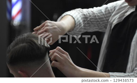 Skilled barber trimming customer's hair using professional scissors and comb, working in contemporary barbershop with vintage pole visible near workspace 129438573