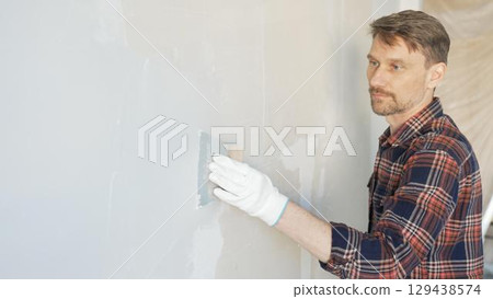 Construction worker man wearing gloves and holding a putty knife is applying plaster on a white wall, performing home renovation and improvement 129438574