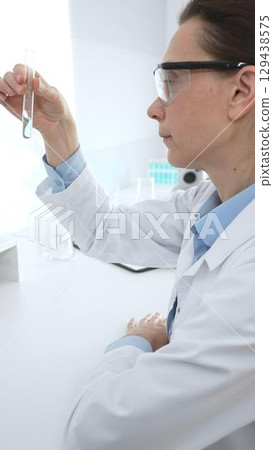 Female researcher examining clear liquid in test tube, sitting near microscopes and lab equipment within sterile scientific workspace. Medicine, healthcare and science concept 129438575