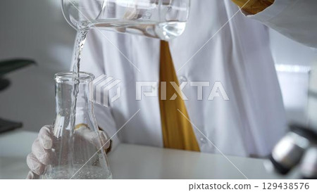 Unknown scientist with a lab coat and white protective gloves is carefully pouring a liquid from a beaker into an Erlenmeyer flask, close up. Science and medicine concept 129438576