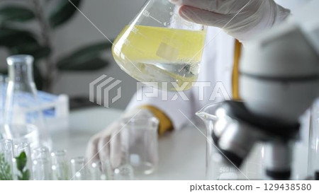 A scientist wearing white protective gloves is shaking up a yellow chemical solution inside an Erlenmeyer flask in a laboratory set, close up. Science and medicine concepts A scientist wearing white protective gloves is shaking up a yellow chemical solution inside an Erlenmeyer flask in a laboratory set, close up. Science and medicine concepts 129438580