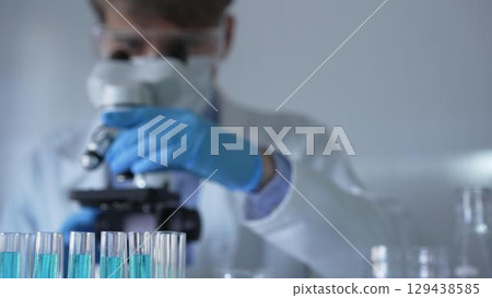 Laboratory tubes are in focus, closeup view. Man scientist researcher with a blue protective gloves is using a microscope at the background. Medicine and science concept 129438585