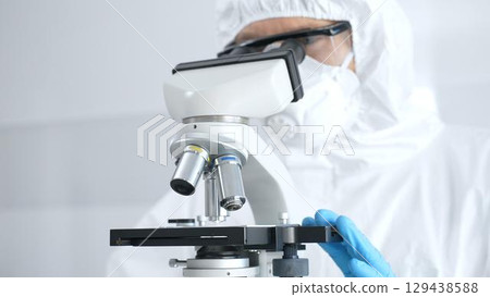 Medical virologist in a hazmat suit, mask, and blue protective gloves, carefully working at a microscope in a laboratory researching viruses, close up. Medicine and science concept Medical virologist in a hazmat suit, mask, and blue protective gloves, carefully working at a microscope in a laboratory researching viruses, close up. Medicine and science concept 129438588