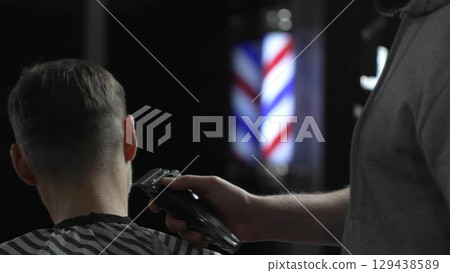 Male professional barber trimming client's hair with electric clipper, vintage illuminated barber pole visible in background, creating contemporary grooming experience Male professional barber trimming client's hair with electric clipper, vintage illuminated barber pole visible in background, creating contemporary grooming experience 129438589