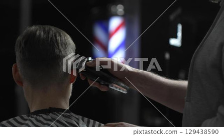 Male professional barber trimming client's hair with electric clipper, vintage illuminated barber pole visible in background, creating contemporary grooming experience Male professional barber trimming client's hair with electric clipper, vintage illuminated barber pole visible in background, creating contemporary grooming experience 129438590