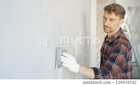 Construction worker man wearing gloves and holding a putty knife is applying plaster on a white wall, performing home renovation and improvement Construction worker man wearing gloves and holding a putty knife is applying plaster on a white wall, performing home renovation and improvement 129438592