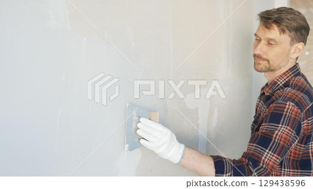 Construction worker man wearing gloves and holding a putty knife is applying plaster on a white wall, performing home renovation and improvement 129438596