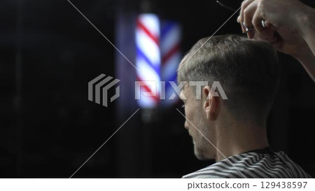 Professional barber cutting hair of male client in barbershop, using scissors and comb, with barber pole in background, creating stylish and modern haircut 129438597