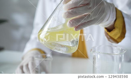 A scientist wearing white protective gloves is shaking up a yellow chemical solution inside an Erlenmeyer flask in a laboratory set, close up. Science and medicine concepts 129438598
