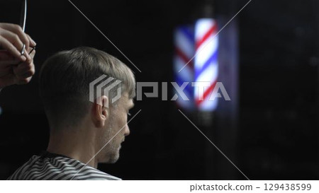 Professional barber grooming male client's hair with scissors, vintage barber pole framing traditional barbershop setting 129438599