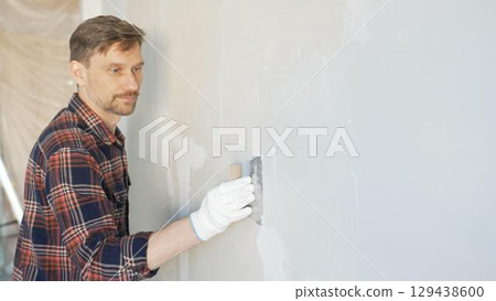 Construction worker man wearing gloves and holding a putty knife is applying plaster on a white wall, performing home renovation and improvement Construction worker man wearing gloves and holding a putty knife is applying plaster on a white wall, performing home renovation and improvement 129438600