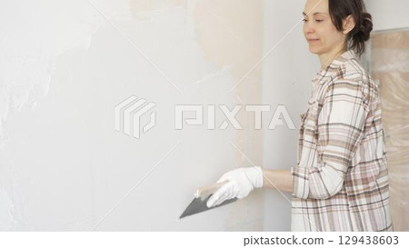 Woman applying plaster to wall with putty knife, wearing protective gloves and creating smooth surface during home renovation work 129438603