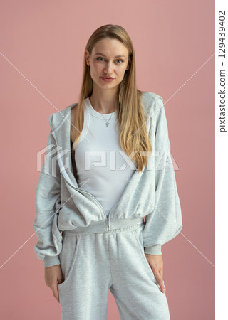 Woman stands against pink background wearing light gray tracksuit 129439402