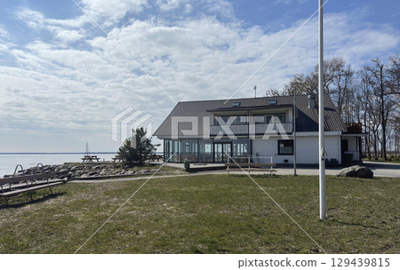 A peaceful lakeside view featuring a two-story white house with large windows, possibly a restaurant or a residential building, overlooking the calm water 129439815