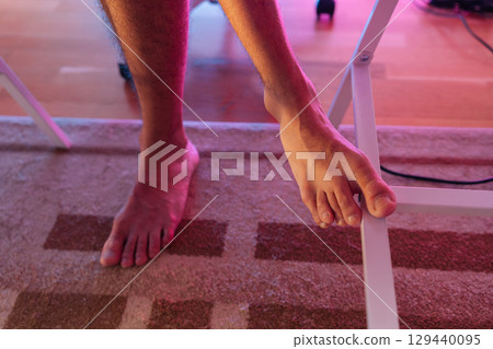 Close-up feet of unrecognizable gamer male resting on white metal chair leg while playing videogames at home, illuminated by pink neon light. Concept of relaxation and comfort during gaming sessions. 129440095