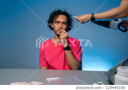 Man looking at camera sitting at table receiving bizarre hair vacuum treatment while applying nasal spray, depicting surreal grooming moment with unexpected comedic twist. Man looking at camera sitting at table receiving bizarre hair vacuum treatment while applying nasal spray, depicting surreal grooming moment with unexpected comedic twist. 129440100