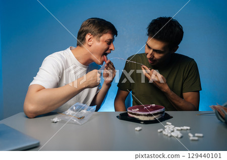 Two friends enjoying drug-fueled party licking icing dripping from cake, with pills and alcohol scattered on table, highlighting dangers of substance abuse. Concept of party with substance abuse. Two friends enjoying drug-fueled party licking icing dripping from cake, with pills and alcohol scattered on table, highlighting dangers of substance abuse. Concept of party with substance abuse. 129440101