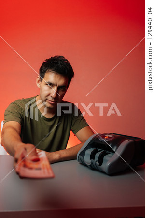 Studio portrait of cashier giving out banknotes looking at camera while positioned next to money counting machine on table, set against vibrant red background, captures moment of financial transaction Studio portrait of cashier giving out banknotes looking at camera while positioned next to money counting machine on table, set against vibrant red background, captures moment of financial transaction 129440104