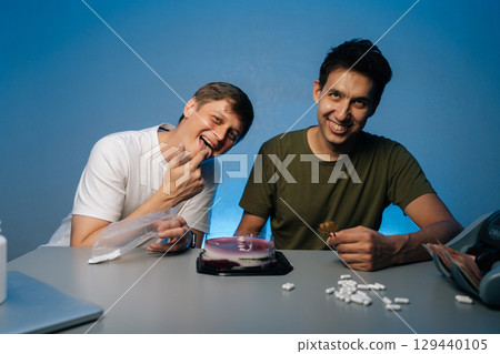 Two cheerful friends sitting at table with drugs, white powder, pills, cake, money in studio with isolated blue background. Concept of party atmosphere with substance abuse. Two cheerful friends sitting at table with drugs, white powder, pills, cake, money in studio with isolated blue background. Concept of party atmosphere with substance abuse. 129440105