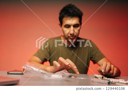 Selective focus shot of drug realer male preparing dose of cocaine or heroin, surrounded by pills, cash and smartphone on red background, highlighting dangers of addiction and illegal drug trade. Selective focus shot of drug realer male preparing dose of cocaine or heroin, surrounded by pills, cash and smartphone on red background, highlighting dangers of addiction and illegal drug trade. 129440106