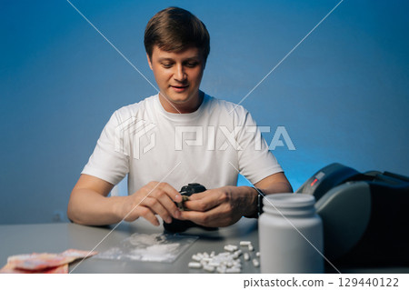 Portrait of young photographer male preparing drugs with banknote, smiling while sitting at table with pills, camera and plastic bag with white powder on blue isolated background. 129440122