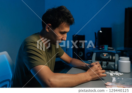 Side of pensive young man preparing dose of drugs sitting at desk cluttered with pills and cash in dark room, studio shot portrait on blue background. Concept of addiction and substance abuse. 129440125
