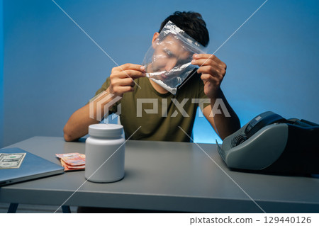Portrait of drug dealer male preparing dose of cocaine or methamphetamine from plastic bag sitting at table with white plastic jar, laptop and counting machine. Concept of drug dealing or addiction. 129440126