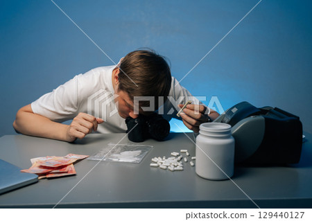 Young photographer male snorting drugs through rolled banknote at workplace, surrounded by drug paraphernalia, pills, money and equipment, highlighting dangers of addiction, studio shot. 129440127