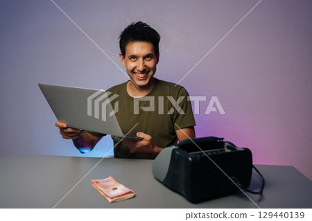 Studio portrait of friendly cashier smiling looking at camera, holding laptop sitting at desk with banknotes and banknote counter machine, suggesting modern approach to finance and money management 129440139