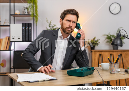 Middle-aged businessman lifts retro wired telephone and makes client call listening closely calmly Middle-aged businessman lifts retro wired telephone and makes client call listening closely calmly 129440167
