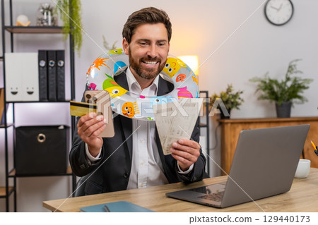 Middle-aged businessman pays for beach holiday tickets smiles closes laptop puts inflatable ring joy Middle-aged businessman pays for beach holiday tickets smiles closes laptop puts inflatable ring joy 129440173