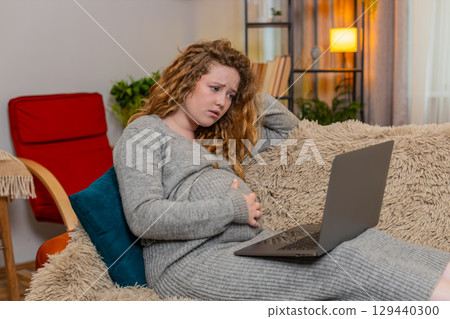 Pregnant woman massaging temples while working on laptop at home, tired stressed expression fatigue 129440300