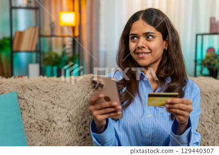 Indian woman smiling while shopping online using smartphone and credit card on home sofa indoors Indian woman smiling while shopping online using smartphone and credit card on home sofa indoors 129440307