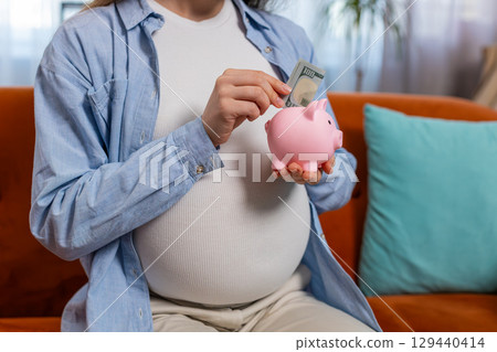 Pregnant woman inserting banknote money cash dollar in piggybank saving for baby's future needs Pregnant woman inserting banknote money cash dollar in piggybank saving for baby's future needs 129440414