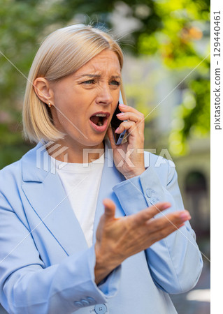 Angry mature Caucasian businesswoman arguing while talking on smartphone standing on city street 129440461