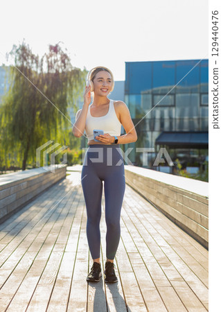 Happy young woman jogger listening to music via headphones and smartphone walking in city park Happy young woman jogger listening to music via headphones and smartphone walking in city park 129440476