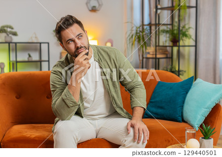 Sad lonely Caucasian young man sitting on sofa at home room looks pensive thinks over life concerns 129440501